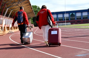 Kobieta i mężczyzna w czerwonych bluzach idą po bieżni stadionu ciągnąc za sobą walizkę i pojemnik.
