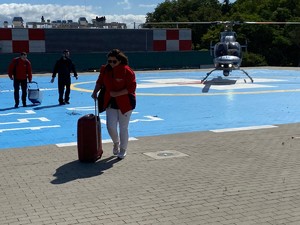 Kobieta w czerwonej bluzie i okularach przeciwsłonecznych z walizką na kółkach wychodzi z lądowiska, za nią mężczyzna w towarzystwie policyjnego pilota, w tle śmigłowiec.