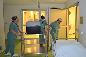 Pracownicy szpitala transportują pojemnik z organem na wózku, drzwi otwiera im osoba w masce na twarzy.