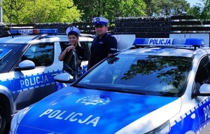 policjantka i policjant ruchu drogowego w czapkach z białymi pokrowcami stoją pomiędzy oznakowanymi radiowozami