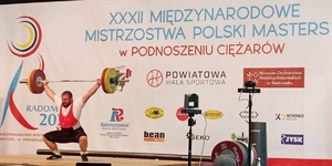 zdjęcie z zawodów w podnoszeniu ciężarów - mężczyzna podnosi sztangę, z tyłu widoczna jest ścianka a nie niej m.in. napis: XXXII Międzynarodowe Mistrzostwa Polski Masters w Radomsku