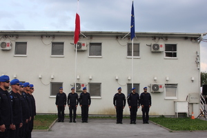 na zdjęciu grupa funkcjonariuszy stoi tyłem do budynku, w tle widać maszty z zawieszonymi flagami Polski i Unii Europejskiej