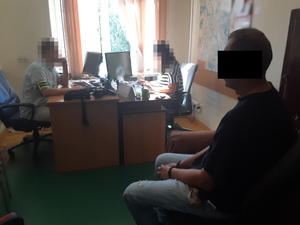 zatrzymany mężczyzna zakuty w kajdankach siedzi na krześle, w tle nieumundurowany policjant i policjantka siedzą naprzeciwko siebie przy biurkach