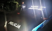 na zdjęciu w nocy stojący radiowóz na światłach, obok stojący policjant, w tle krzewy