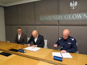 Dwaj mężczyźni w garniturach i oficer policji siedzą za stołem. Drugi od lewej mężczyzna w garniturze podpisuje porozumienie oraz siedzący dalej oficer Policji w mundurze również podpisuje porozumienie.