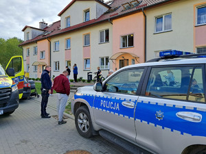na zdjęciu obok radiowozu policyjnego stoi funkcjonariusz i mężczyzna, obok policjanta stoi zaparkowana żółta karetka pogotowia, w tle budynek mieszkalny