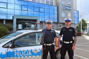 dwóch umundurowanych policjantów stoi przy radiowozie, w tle budynek komendy policji