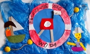 na środku pracy plastycznej widać koło ratunkowe na niebieskim tle z napisem BEZPIECZNIE NAD WODĄ, po dwóch stronach koła naklejono kolorowe postacie na łódkach