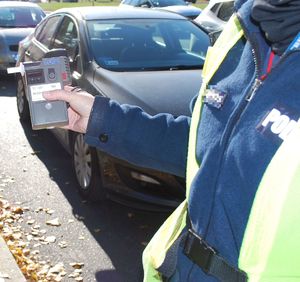 Na zdjęciu umundurowana policjantka ruchu drogowego w odblaskowej kamizelce trzyma w wyciągniętej dłoni urządzenie do kontroli stanu trzeźwości. W tel stojące przy krawędzi jezdni samochody