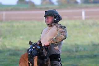 Policyjny przewodnik psa w umundurowaniu taktycznym i hełmie na głowie przytrzymuje swojego podopiecznego, pies ma na głowie specjalne gogle i kaganiec na pysku.