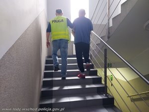 Policjant w żółtej kamizelce prowadzi po schodach zatrzymanego mężczyznę