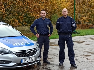 Dwaj policjanci stojący obok radiowozu w terenie zielonym