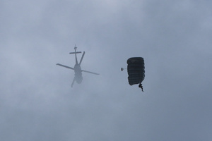 Sylwetka śmigłowca ginącego w chmurach, obok sylwetka opadającego spadochroniarza.