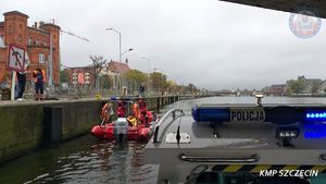widok z policyjnej motorówki z wody na łódź WOPR zacumowanej przy nabrzeżu, w której siedzi mężczyzna owinięty kocem termicznym, przy nim stoją dwaj ratownicy, na nabrzeżu stoją trzy osoby