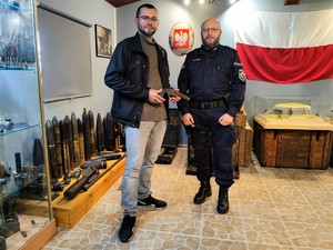 Zdjęcie kolorowe przedstawia umundurowanego policjanta obok mężczyzny, który trzyma w rękach zabytkowy pistolet
