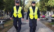 dwaj umundurowani policjanci w żółtych odblaskowych kamizelkach idą alejka na cmentarzu - widok z przodu