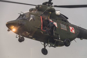 Mężczyzna wiszący przy burcie ciemno-zielonego helikoptera.