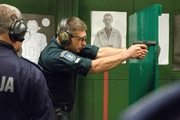 Policjant litewski strzela zza drewnianej osłony stojąc profilem do obiektywu aparatu. Broń zwrócona w prawą stronę.