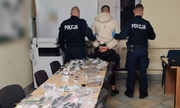 dwaj policjanci z zatrzymanym mężczyzną w pomieszczeniu, na długim stole leżą zabezpieczone narkotyki w woreczkach