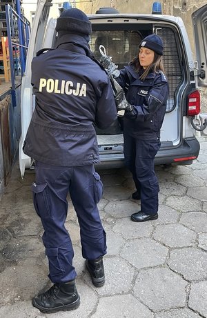 Umundurowany policjant przekazuje worki, które podaje mu policjantka z bagażnika otwartego radiowozu