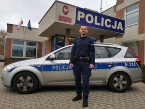 umundurowany policjant stoi przy radiowozie, w tle budynek jednostki Policji