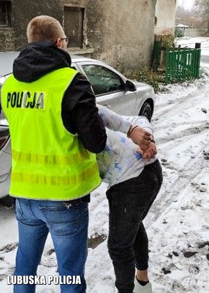 policjant w żółtej kamizelce z napisem Policja na plecach, prowadzi do samochodu zatrzymanego mężczyznę