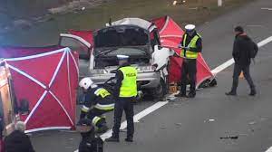 policjanci pracują na miejscu wypadku drogowego. Na jezdni widoczny jest rozbity samochód i rozstawione dwa czerwone namioty