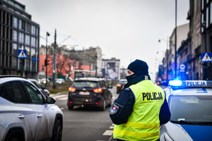 ulica, policjant stoi tyłem w kamizelce odblaskowej z napisem Policja, po prawej stronie stoi radiowóz na sygnałach świetlnych