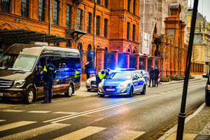 ulica, po prawej stronie są zaparkowane samochody, na prawym pasie stoi radiowóz na sygnałach świetlnych, przy radiowozie policjant pokazuje palcem kierunek, drugi policjant stoi przy brązowym samochodzie zaparkowanym na poboczu