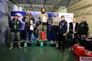 trzej mężczyźni z pucharami, medalami i nagrodami na podium, wokół nich stoją trzej mężczyźni i kobieta