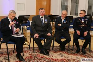 trzej mężczyźni w garniturach i jeden w mundurze przedwojennego policjanta siedzą na krzesłach, jeden z mężczyzn trzyma w reku mikrofon