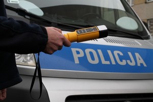 Policjant trzymający w ręku urządzenie do badania stanu trzeźwości na tle przedniej części radiowozu z białym napisem Policja na niebieskim pasie