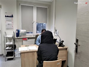 zdjęcie przedstawia moment przesłuchania podejrzanej w jednym z pomieszczeń komisariatu. Zatrzymana kobieta ma na sobie czarną kurtkę