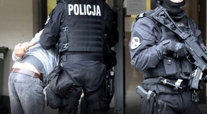 umundurowany policjant prowadzi zatrzymanego - widok z tyłu, obok, przodem do fotografującego stoi policjant z długą bronią
