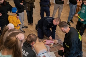 Policjanci i strażacy pokazują grupie dzieci jak udzielić pierwszej pomocy małemu dziecku.