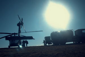 Śmigłowiec stoi na trawiastym lądowisku, obok niego kilka osób oraz samochód ciężarowy z cysterną (zdjęcie wykonane pod słońce).