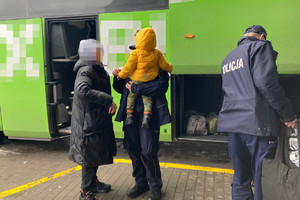 przed autobusem stoi kobieta  z umundurowaną policjantką, która trzyma na reku dziecko, a z boku widać policjanta niosącego bagaże