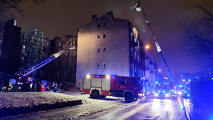 Zdjęcie przedstawia wozy strażackie, które znajdują się po obu stronach budynku. Oba mają uruchomione niebieskie sygnały świetlne