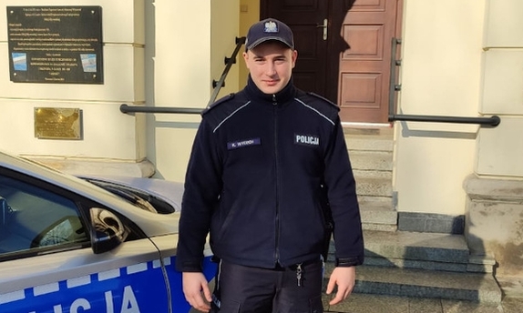 Policjant stoi przed budynkiem Komendy Stołecznej Policji obok radiowozu