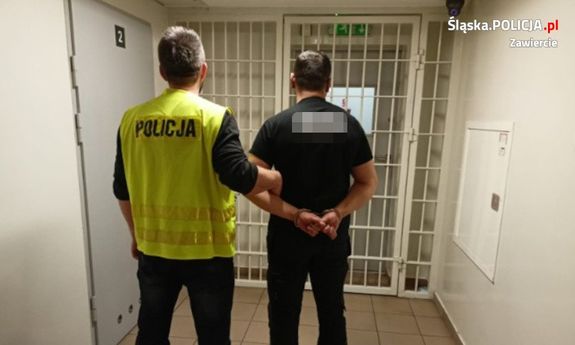 Na kolorowym zdjęciu widoczny policjant w kamizelce odblaskowej z napisem: POLICJA, który prowadzi zatrzymanego mężczyznę przez korytarz Pomieszczeń dla osób zatrzymanych. Mężczyzna ma założone kajdanki na ręce trzymane z tyłu