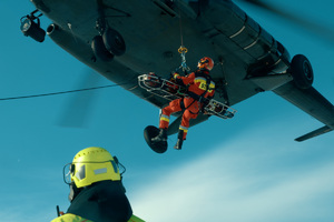 Mężczyzna w żółtym kasku obserwuje wciąganie strażaka ze rannym na specjalnych noszach do śmigłowca.