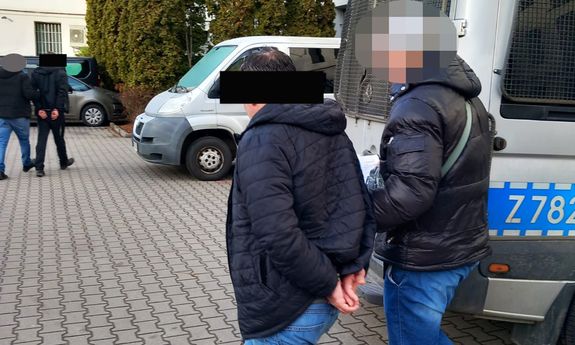 Policjant prowadzi zatrzymanego mężczyznę, który ma założone kajdanki na ręce trzymane z tyłu