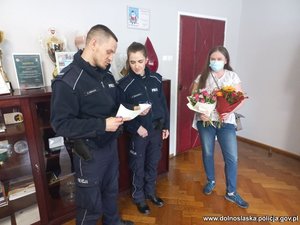 policjant wraz z policjantką czytają podziękowania. Obok stoi mama dziewczynki wraz z bukietem kwiatów, której córeczka te podziękowania napisała
