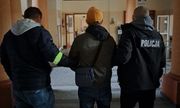 dwaj ubrani po cywilnemu policjanci  w kurtkach z napisem Policja na plecach, prowadzą zatrzymanego