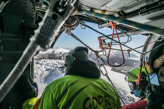 Ratownicy GOPR na pokładzie policyjnego śmigłowca, przez otwarte drzwi widać ośnieżone szczyty górskie.
