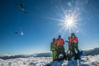 Grupa ratowników górskich obserwuje policyjny śmigłowiec pod którym zwisają na linach dwie osoby.