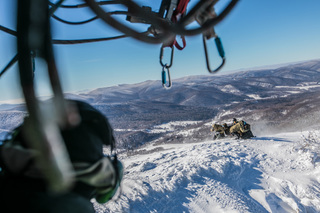 Mężczyzna w hełmie obserwuje z pokładu śmigłowca kilku mężczyzn na zaśnieżonym stoku.