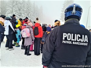 grupa osób i narciarzy na stoku, z boku widać tyłem ustawionego policjanta, który ma na plecach napis: Policja patrol narciarski