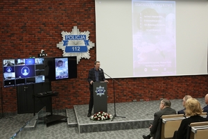 Mężczyzna przemawia stojąc przy mównicy, w tle na ścianie gwiazda policyjna, po lewej stronie zestaw do wideokonferencji