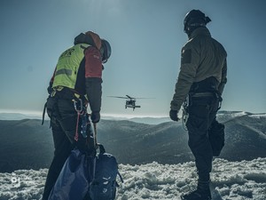 Goprowiec i funkcjonariusz BOA stoją na ośnieżonym stoku, w kierunku mężczyzn leci policyjny Black Hawk, tło zdjęcia stanowią pokryte śniegiem szczyty górskie.